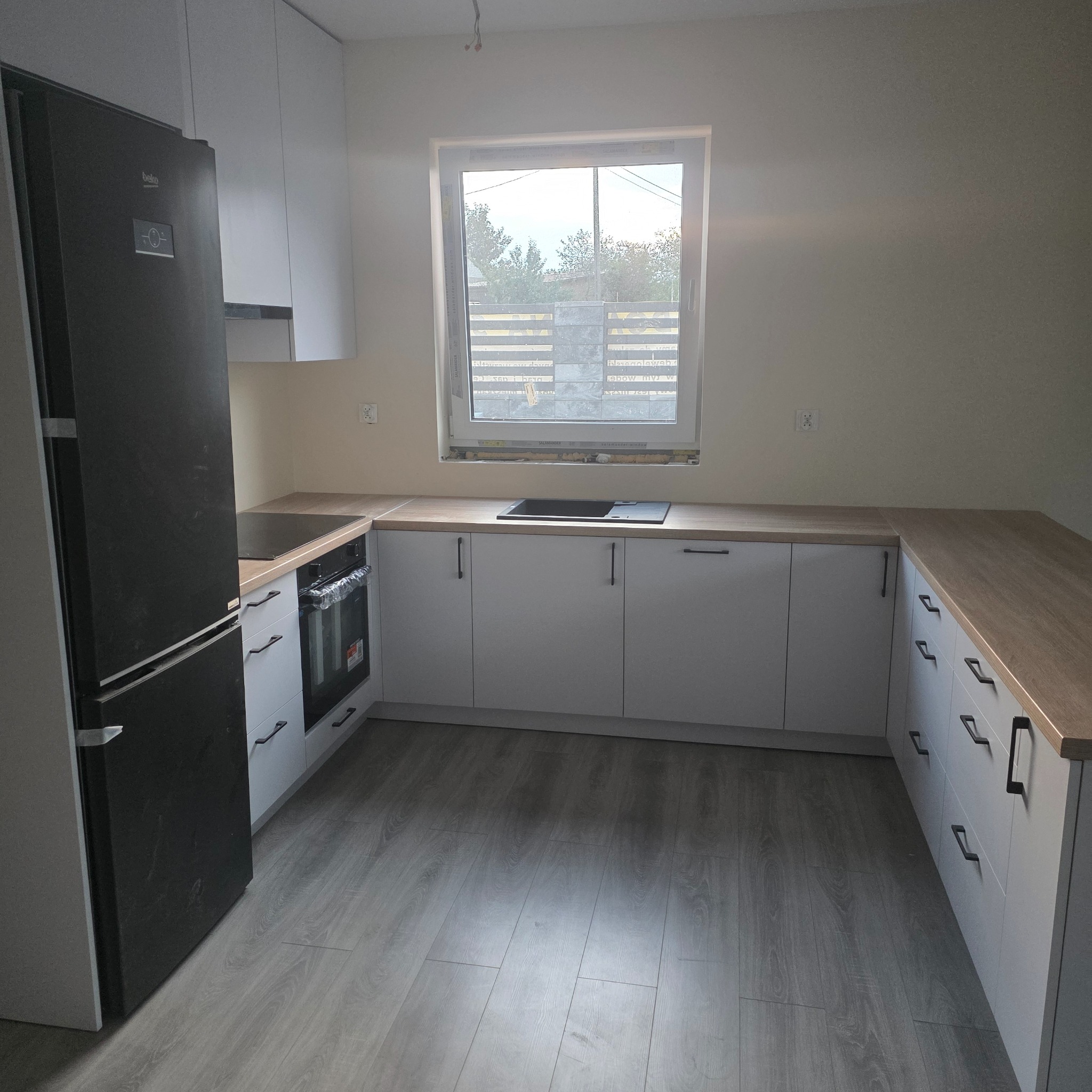 Jasna kuchnia z drewnianym blatem i czarnymi uchwytami, widok na okno z żaluzjami. Nowoczesny design, minimalistyczne wykończenie, szare panele podłogowe.