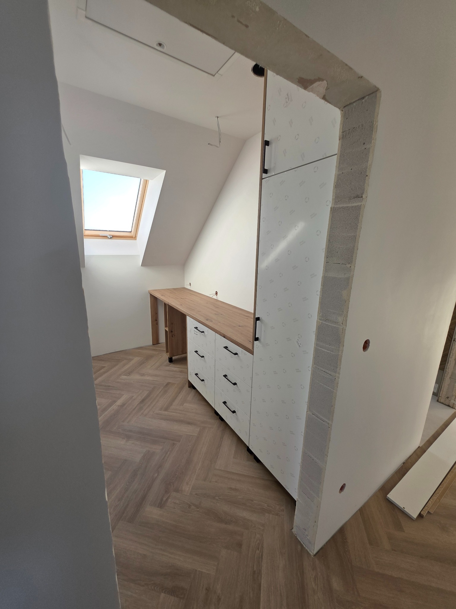 Szafa wnękowa z białymi frontami i czarnymi uchwytami, drewniany blat biurka w skosie poddasza, widok zza surowego otworu drzwiowego.
