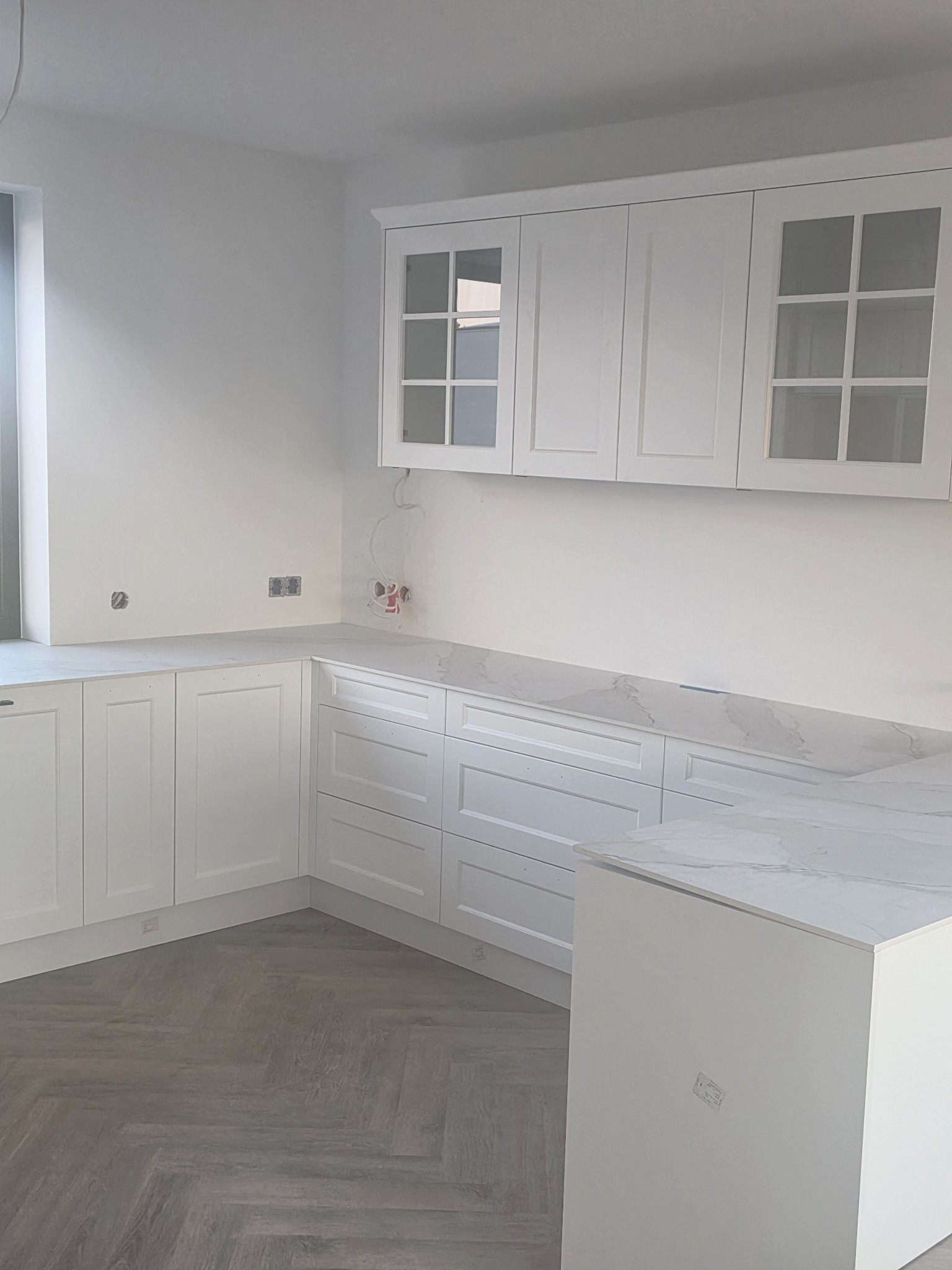 Jasna kuchnia z białymi frontami i marmurowym blatem, widok na szafki wiszące z przeszklonymi drzwiczkami i podłogę ułożoną w jodełkę. Widoczne gniazdka elektryczne.