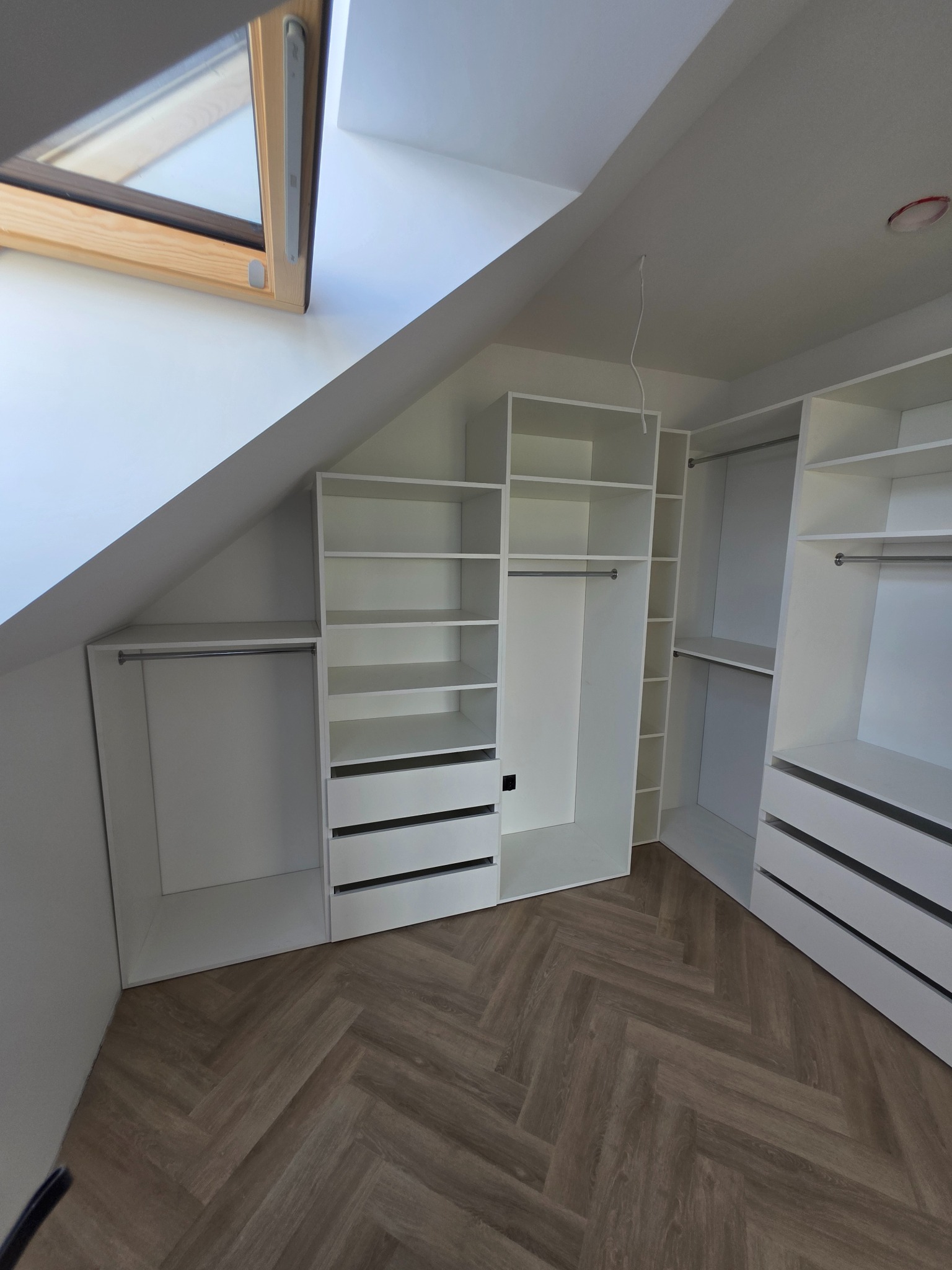 Biała szafa wnękowa na poddaszu z drewnianą podłogą w jodełkę. Widoczne półki, szuflady i drążki na ubrania. Okno dachowe w tle.