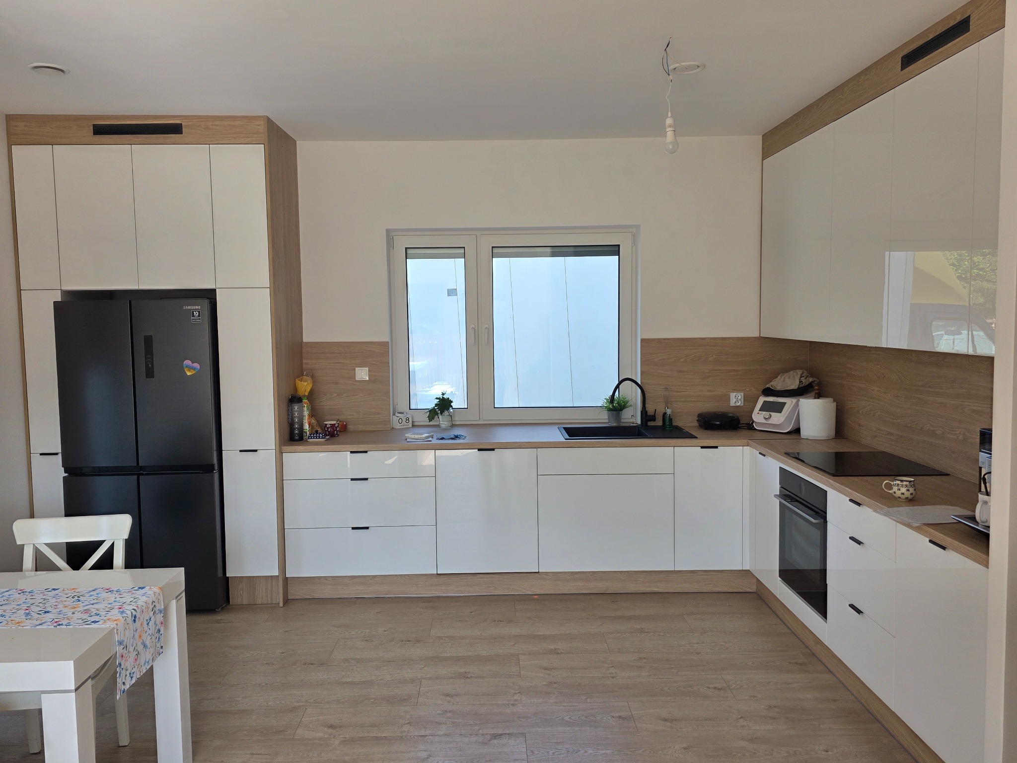 Jasna kuchnia z białymi frontami i drewnianymi akcentami. Czarna lodówka side-by-side. Uwagę zwraca minimalistyczny design i brak górnych szafek.