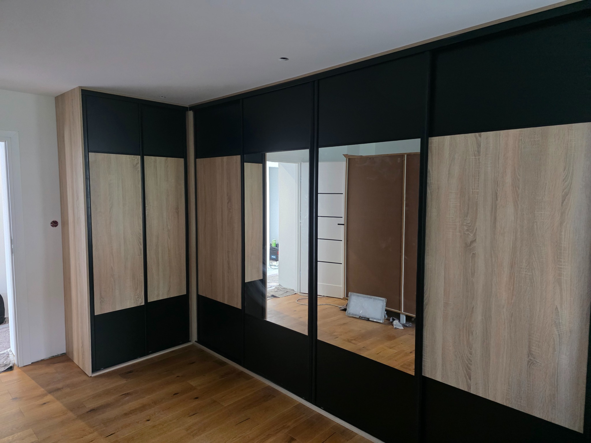 Nowoczesna szafa wnękowa z przesuwnymi drzwiami, łącząca drewnopodobny dekor i czarny mat. Lustrzane panele odbijają wnętrze pokoju, podkreślając przestronność.