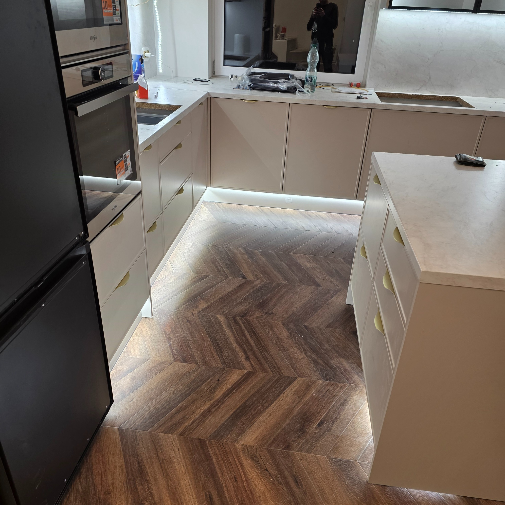 Jasna kuchnia z wyspą i drewnianą podłogą w jodełkę. Podświetlenie LED pod szafkami dodaje ciepła. Widoczny sprzęt AGD i elementy wykończenia.