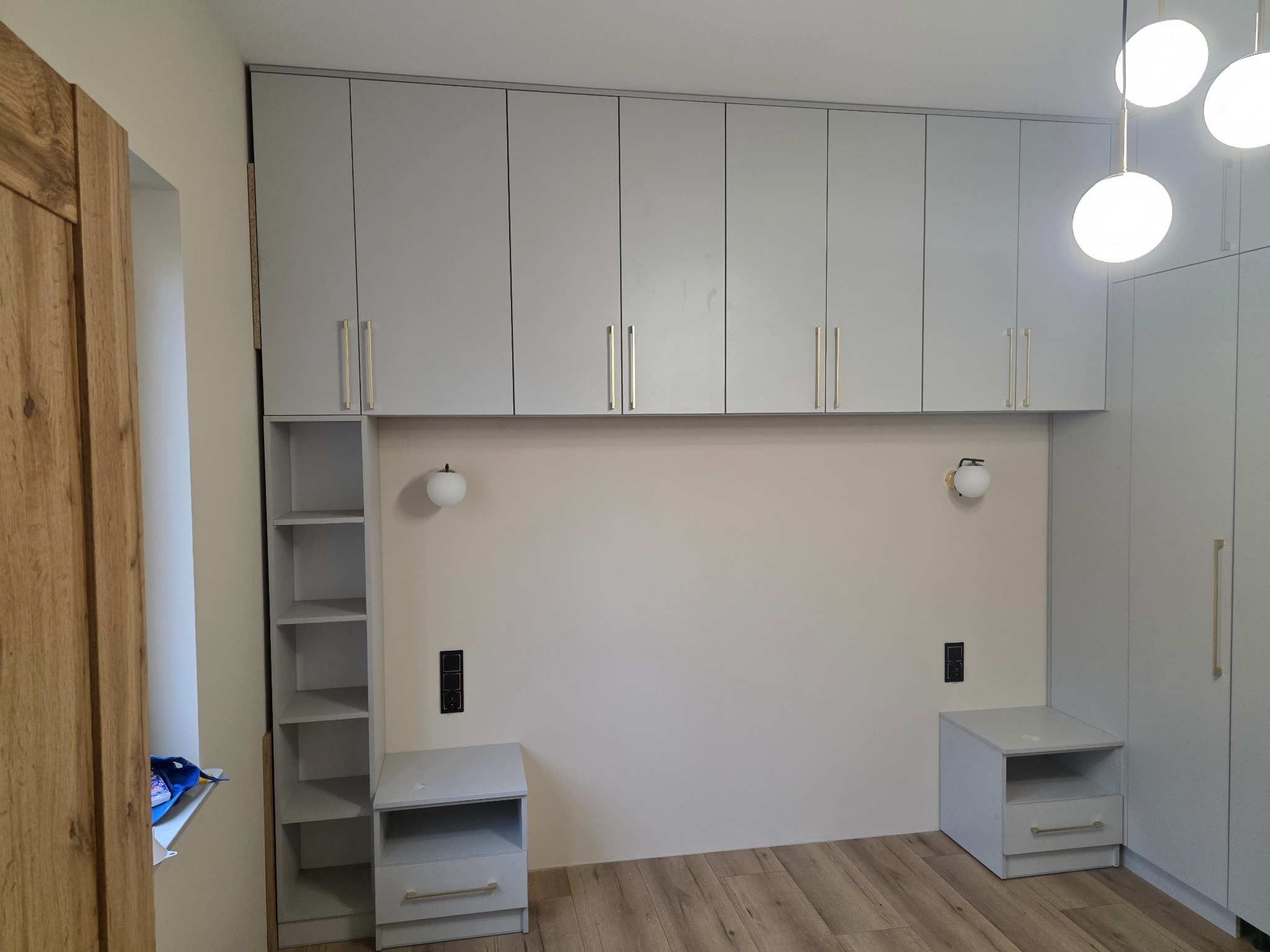 Jasnoszara szafa na wymiar z górnymi szafkami i otwartymi półkami, wkomponowana w aranżację sypialni z szafkami nocnymi i lampkami ściennymi.