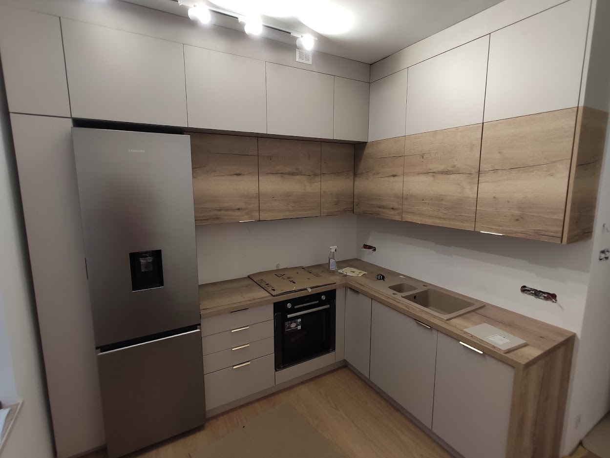 Nowoczesna kuchnia z szarymi szafkami dolnymi i drewnopodobnymi górnymi, srebrna lodówka Samsung, jasne oświetlenie sufitowe, minimalistyczny design.