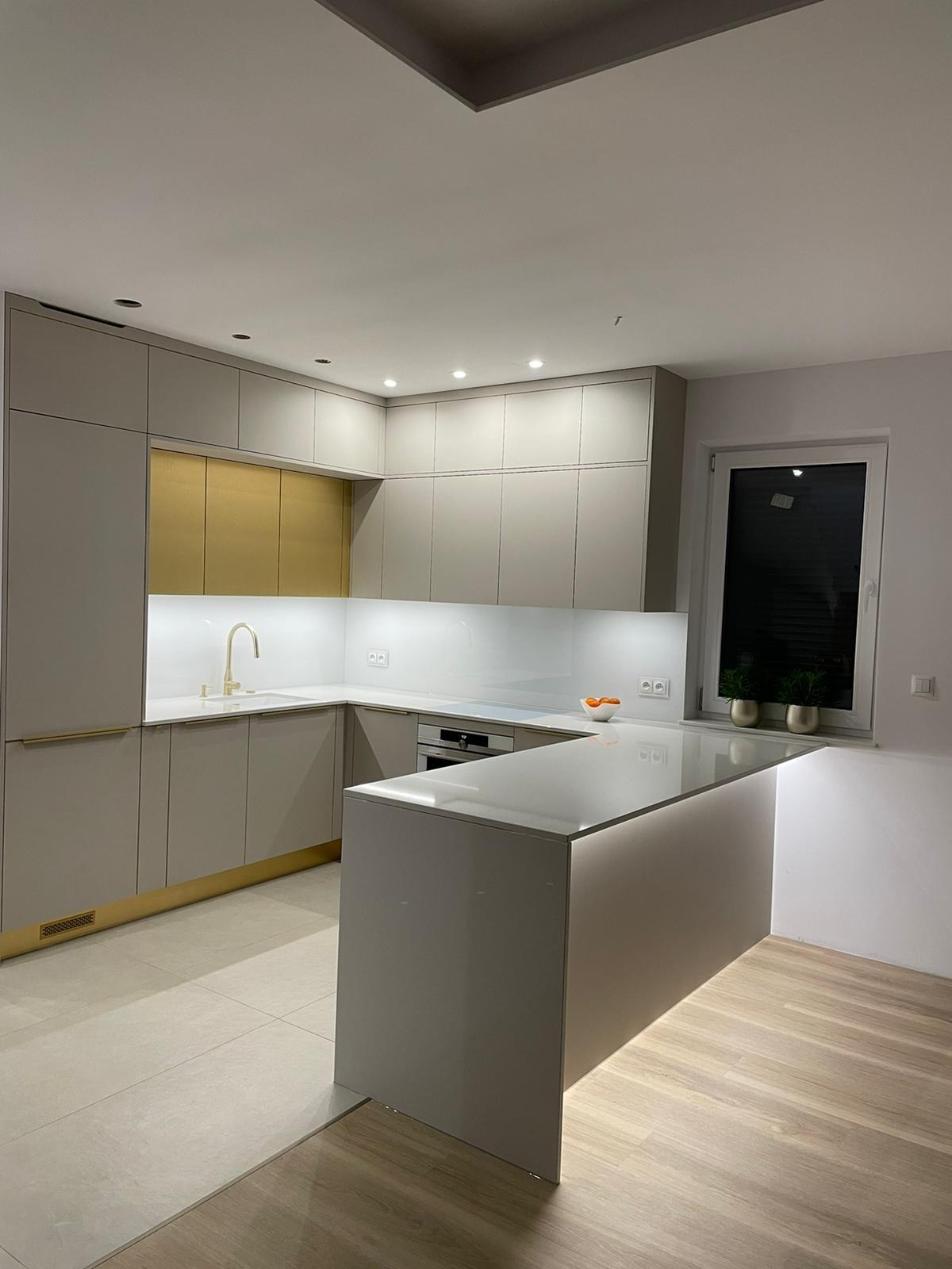 Nowoczesna kuchnia w odcieniach szarości z złotymi akcentami, oświetleniem LED pod wyspą i minimalistycznym designem. Widok na okno z roślinami w tle.