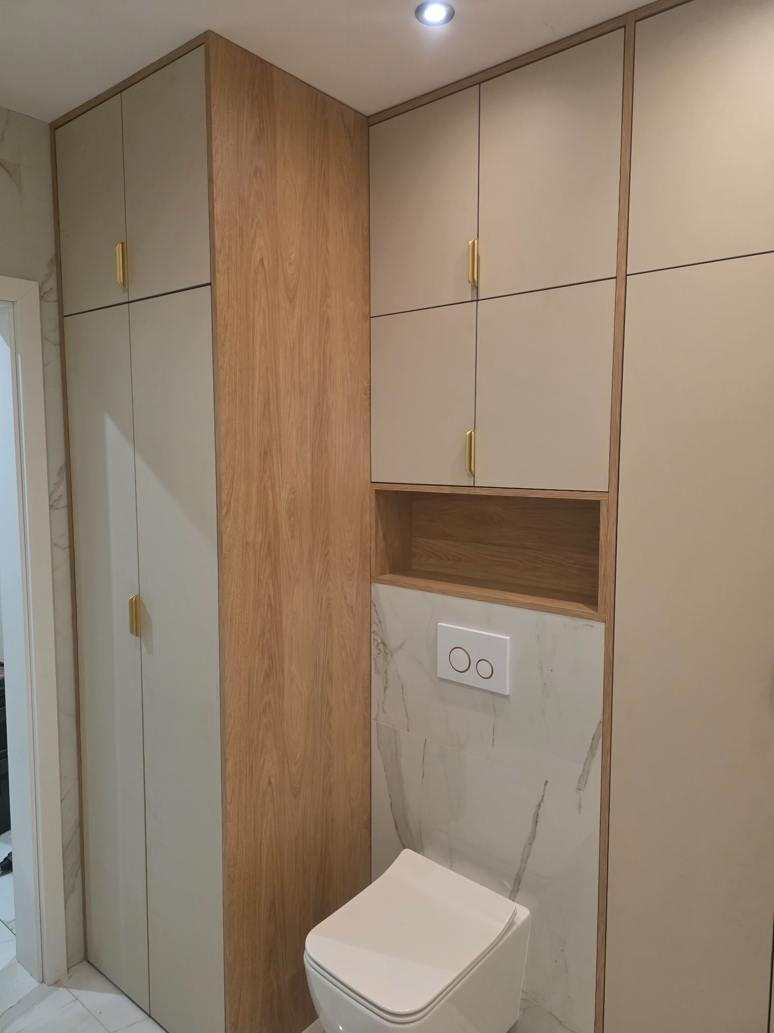 Nowoczesna zabudowa toalety z szafkami w odcieniach beżu i drewna, z minimalistycznymi złotymi uchwytami. Marmurowe płytki i podwieszana miska WC.