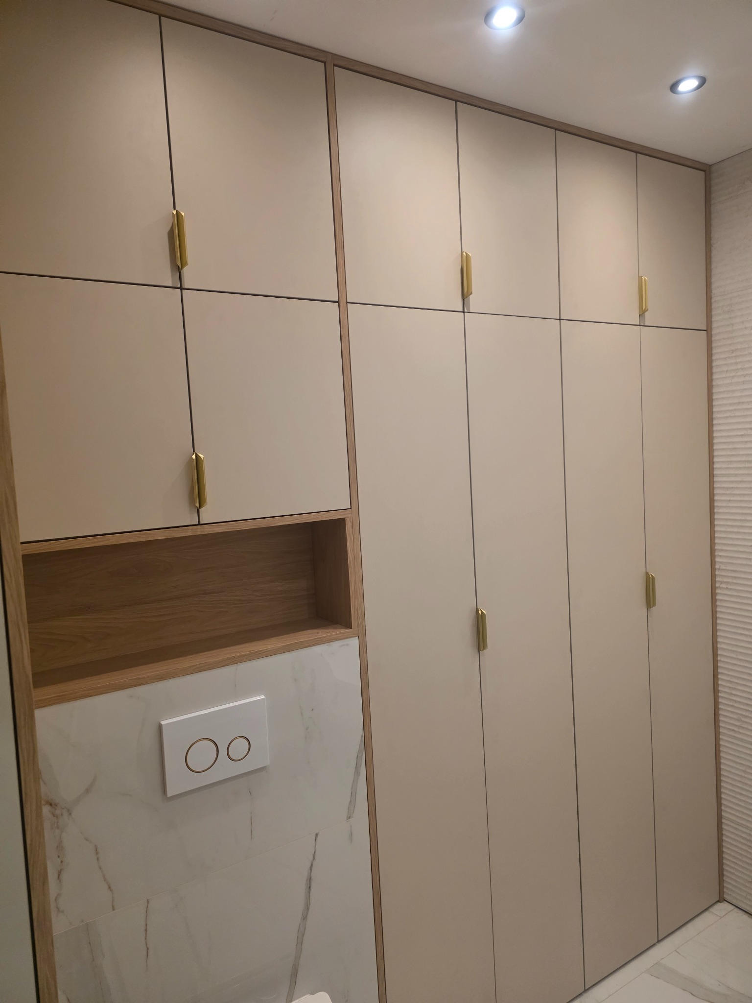 Beżowa szafa wnękowa z złotymi uchwytami, wkomponowana w aranżację łazienki z marmurową ścianą i drewnianymi półkami. Oświetlenie punktowe.
