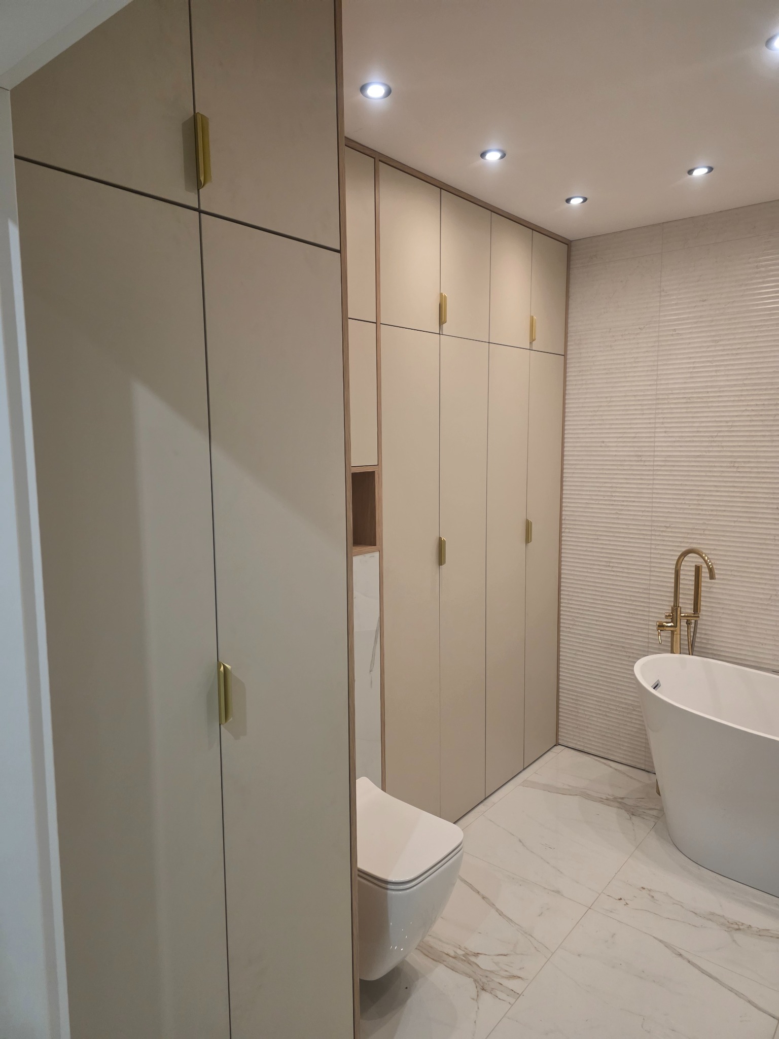 Jasne szafy wnękowe z złotymi uchwytami w nowoczesnej łazience z wanną i toaletą. Minimalistyczny design, marmurowe płytki podłogowe.