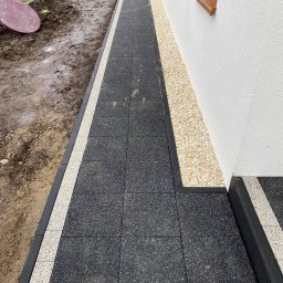 Mateusz Buschan - Ścieżka z ciemnej kostki brukowej obramowana jaśniejszą kostką, biegnąca wzdłuż białej ściany budynku z brązowym parapetem i rabatą z jasnych kamyków.