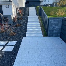 Mateusz Buschan - Nowoczesne podejście do budynku z jasnoszarej kostki brukowej, schody z barierkami, czarny żwir i ozdobne trawy. W tle budynek z numerem 34 i dwa psy na trawniku.