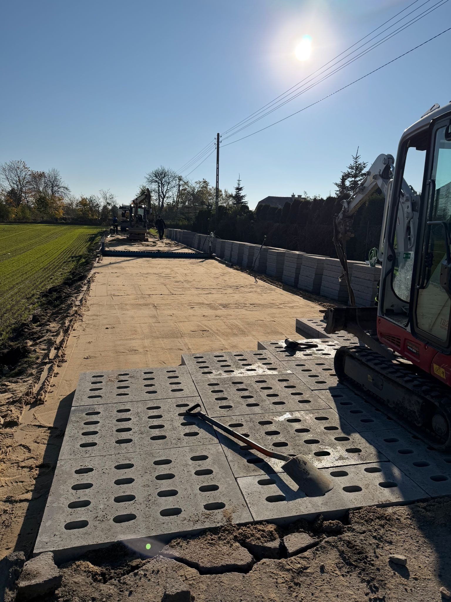 Przygotowanie podłoża z użyciem ażurowych płyt betonowych i minikoparki. Widoczna łopata i ułożone płyty. W tle teren zielony i maszyny budowlane.