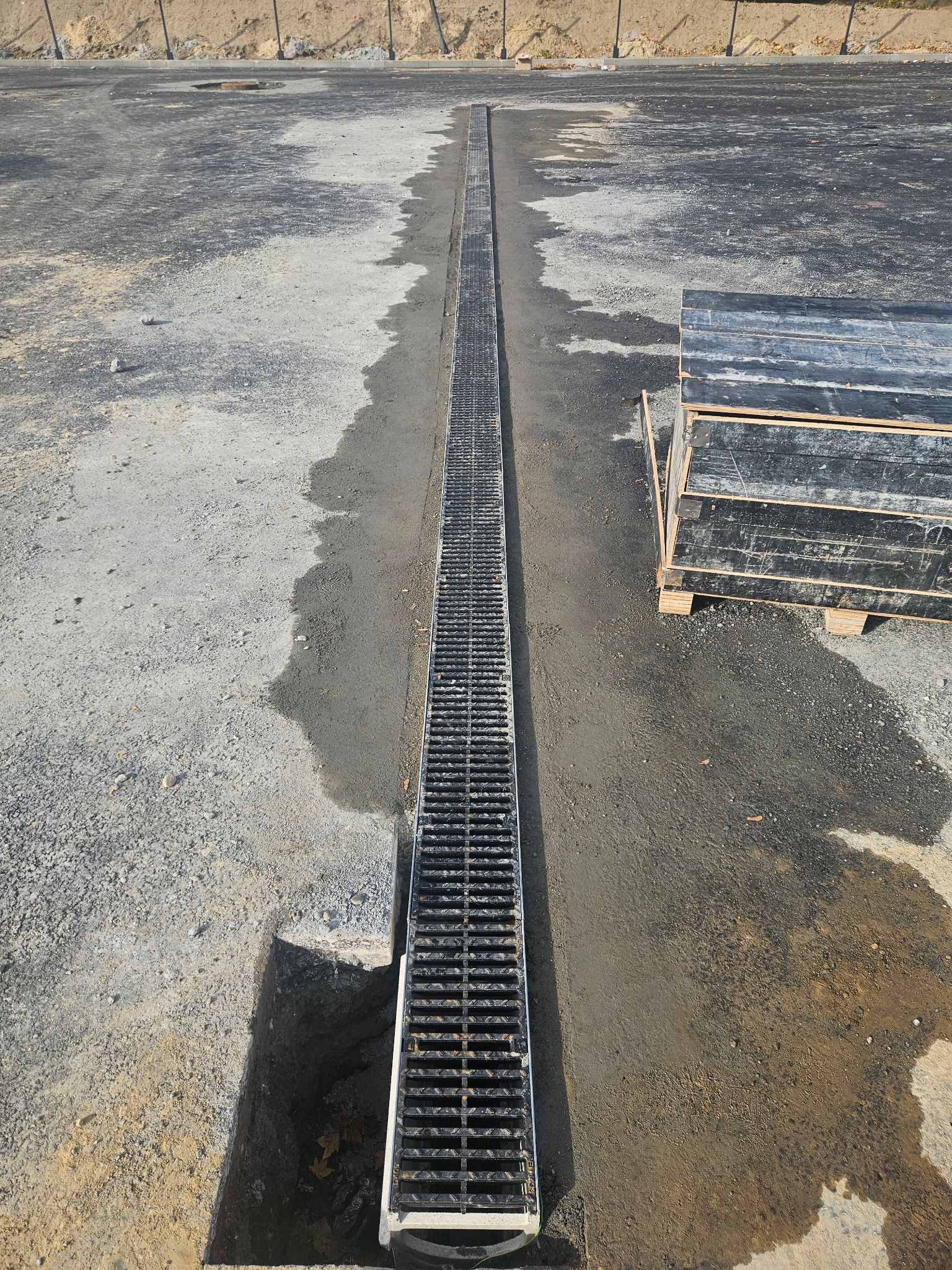Długi, metalowy ruszt drenażowy wkopany w ziemię, biegnący przez utwardzony teren. Obok stos płyt na palecie. Widoczny wykop pod instalację drenażu.