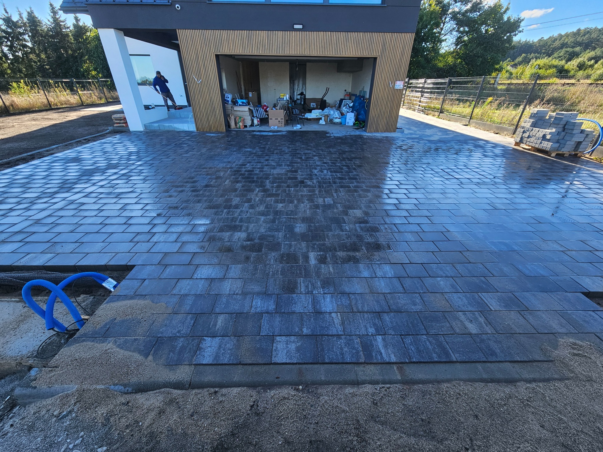 Układanie grafitowej kostki brukowej na podjeździe nowoczesnego domu. Widoczne niebieskie rury drenażowe i paleta z kostką. Prace w toku, częściowo ułożona kostka.