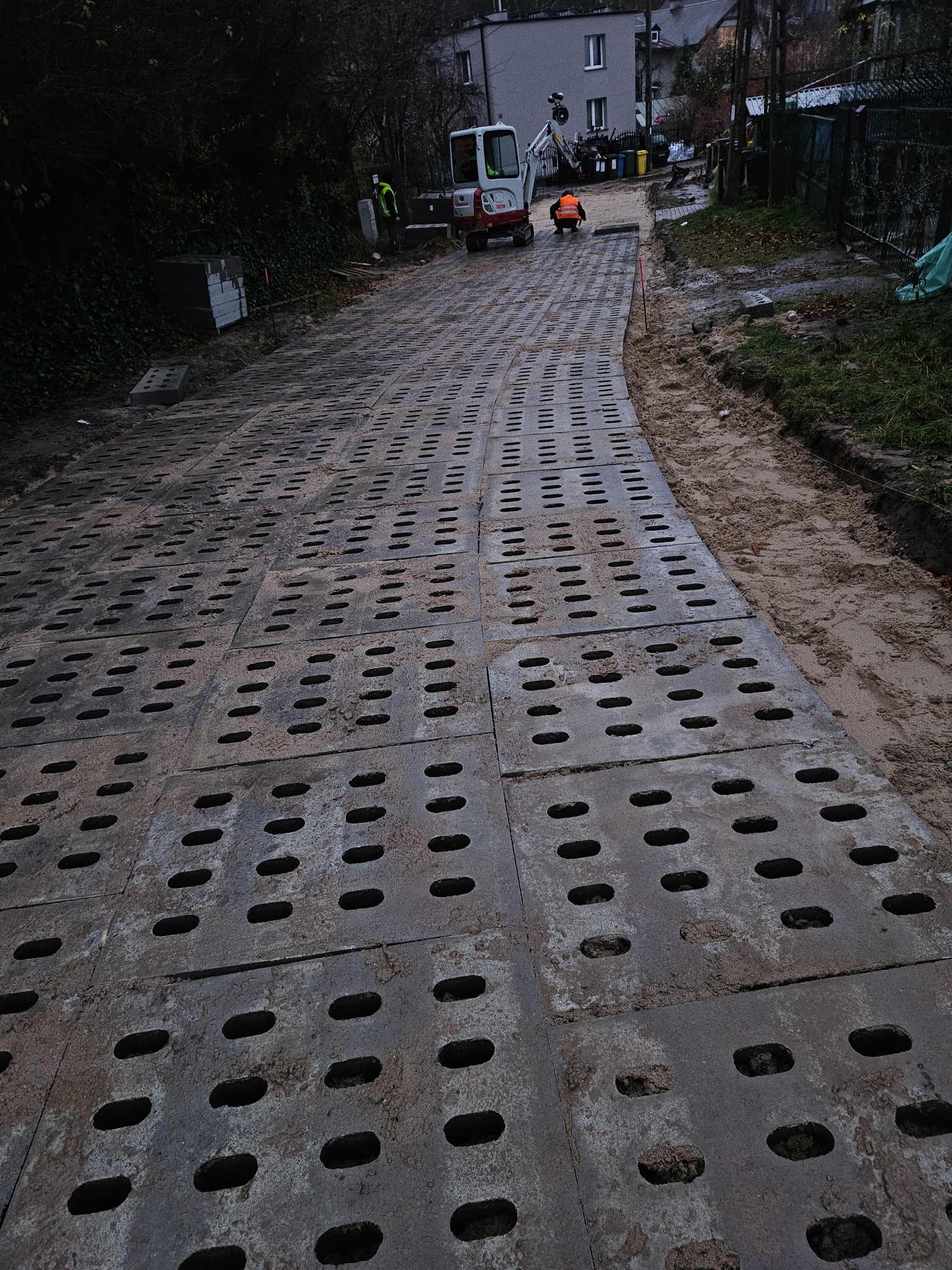 Układanie ażurowych płyt betonowych na drodze gruntowej. W tle minikoparka i pracownicy w odblaskowych ubraniach. Widok z perspektywy ułożonych płyt.