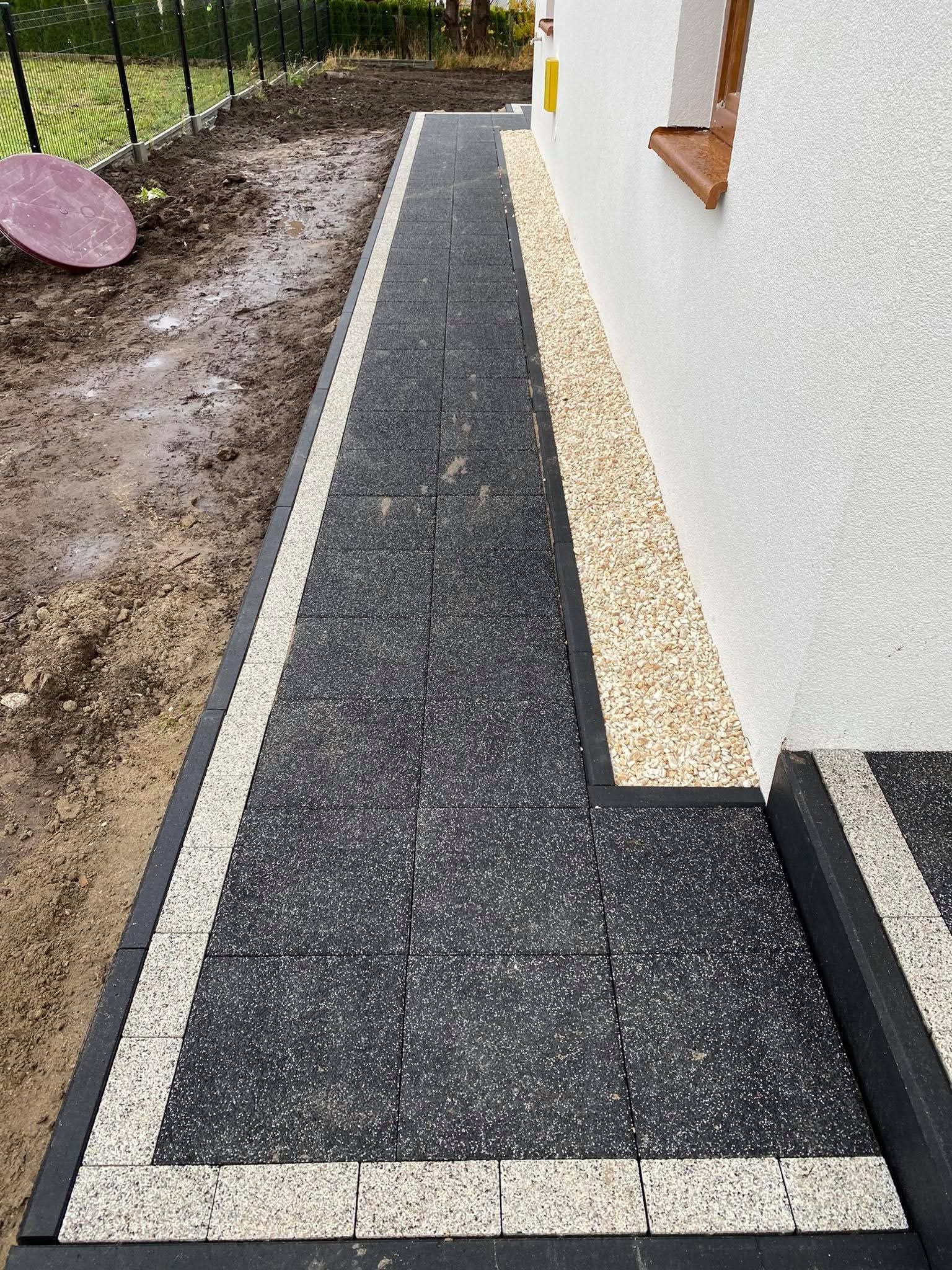 Ścieżka z ciemnej kostki brukowej obramowana jaśniejszą kostką, biegnąca wzdłuż białej ściany budynku z brązowym parapetem i rabatą z jasnych kamyków.