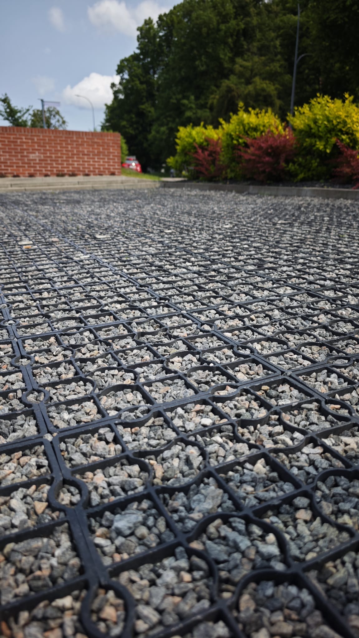 Utwardzona powierzchnia z plastikowej kratki wypełnionej drobnym żwirem, w tle ceglany murek i zieleń. Widok z niskiej perspektywy.
