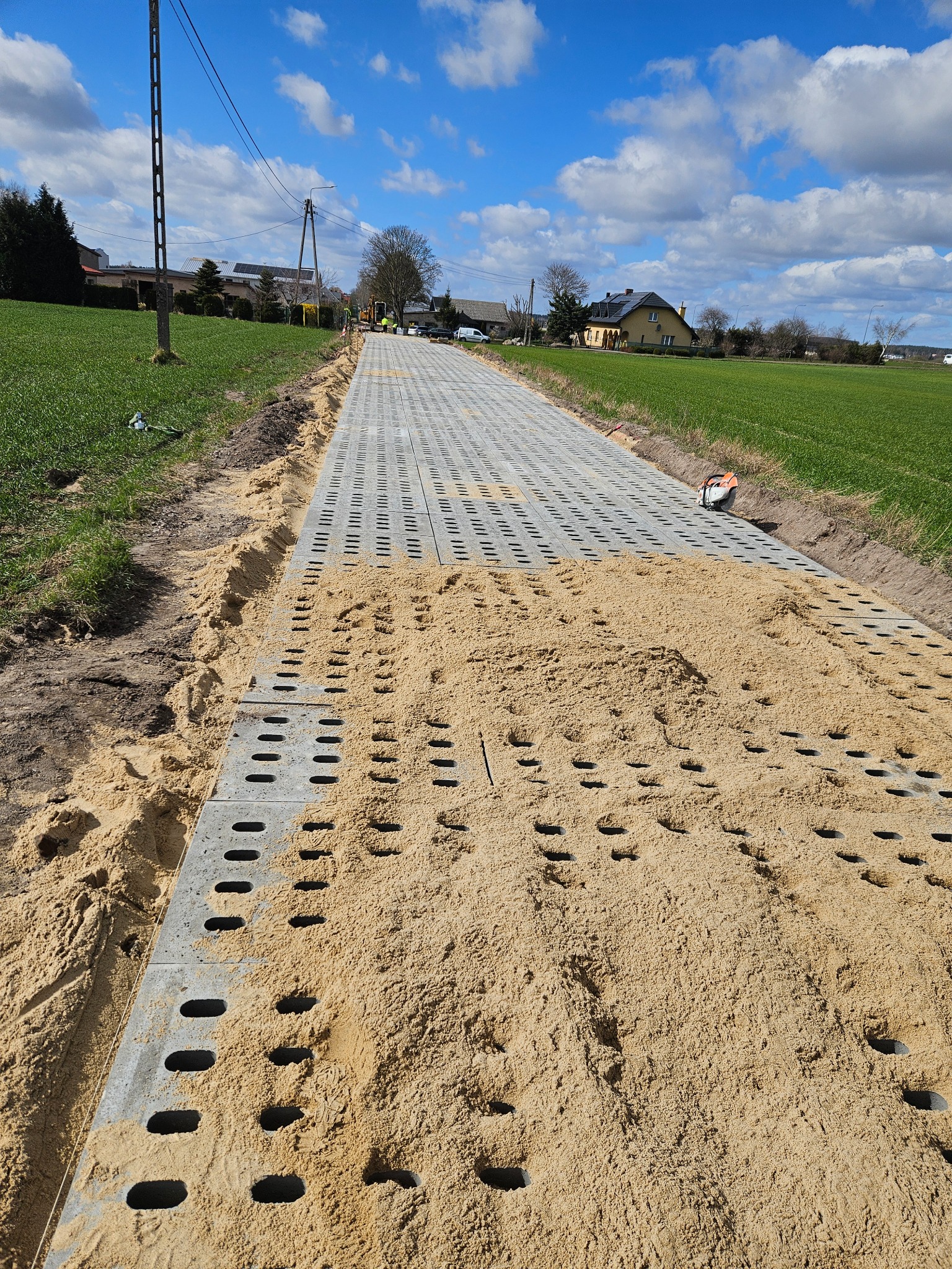 Układanie ażurowych płyt betonowych na piaskowym podłożu, tworzących drogę na tle zieleni i błękitnego nieba. Widoczne narzędzie do cięcia płyt.