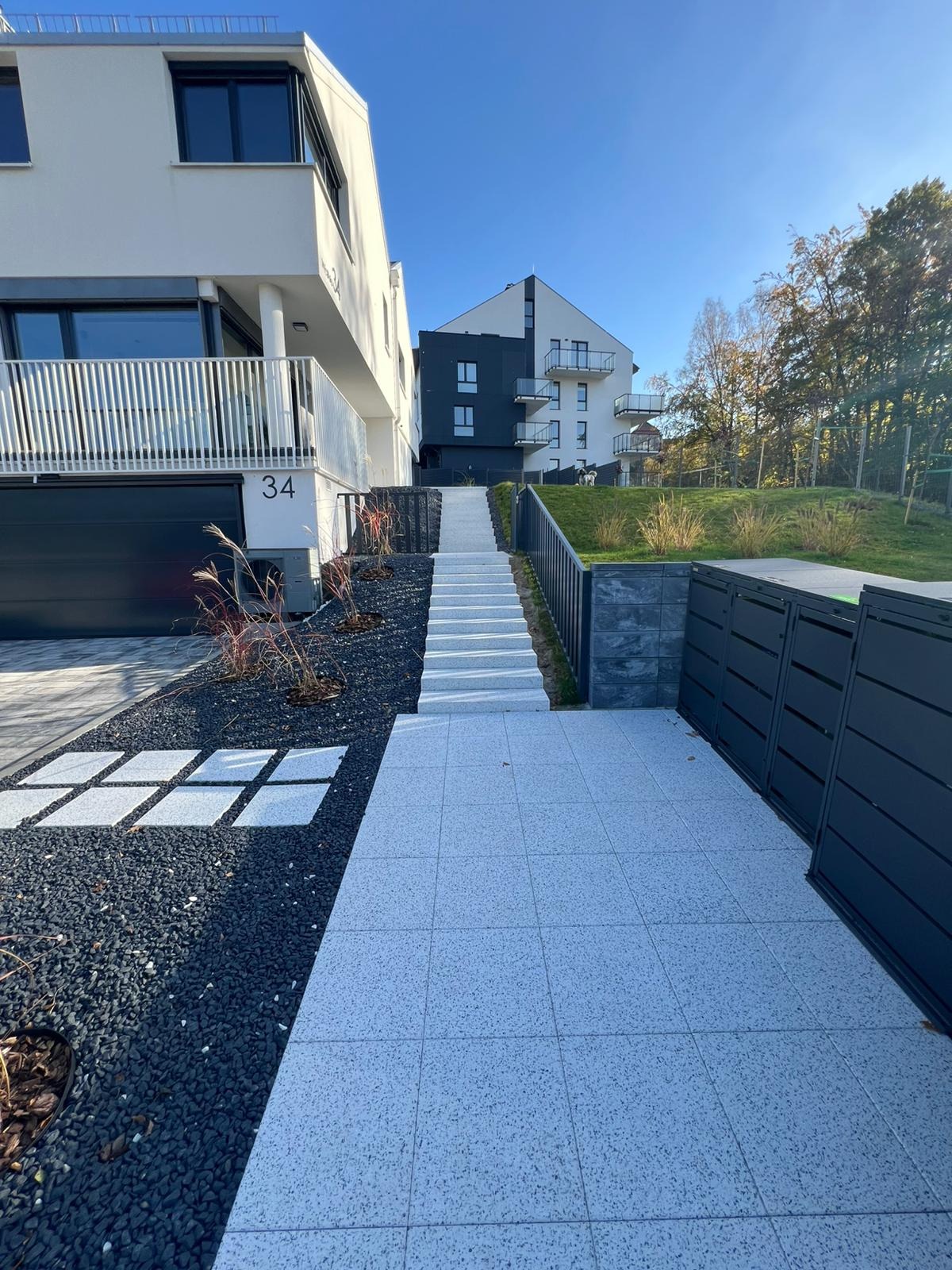 Nowoczesna posesja z jasnoszarą kostką brukową, czarnym grysem i schodami prowadzącymi do budynku. Minimalistyczny design, eleganckie wykończenie.