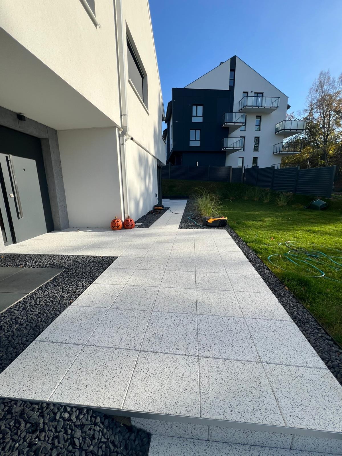 Jasnoszara kostka brukowa ułożona na ścieżce prowadzącej do nowoczesnego domu. Po bokach czarny żwir i trawnik. Dekoracje halloweenowe przy wejściu.