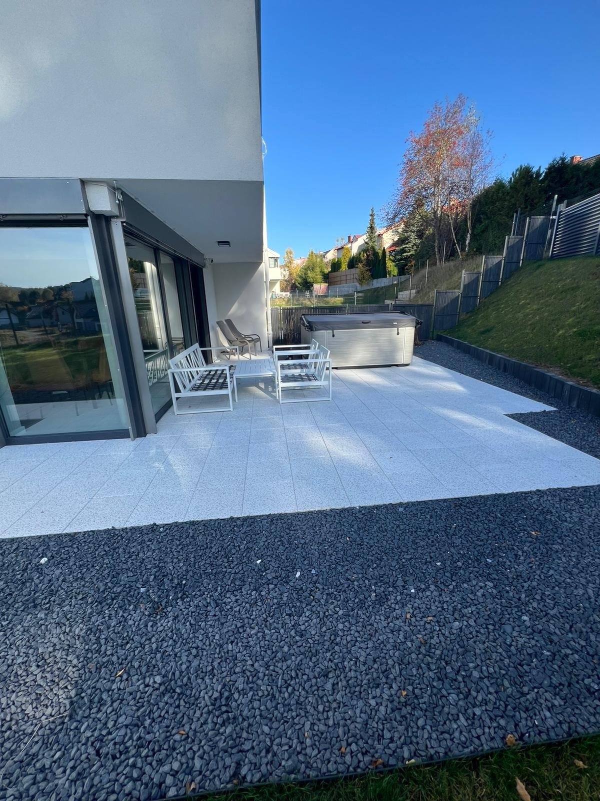 Nowoczesny taras z jasnoszarej kostki, obrzeża z ciemnego grysu, białe meble ogrodowe i jacuzzi na tle domu z dużymi oknami, błękitne niebo.