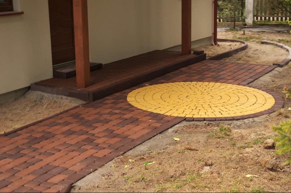 Ułożona z kostki brukowej ścieżka prowadząca do domu, z okrągłym elementem w kolorze żółtym. Kostka w odcieniach brązu i czerwieni. Widoczny fragment elewacji budynku.
