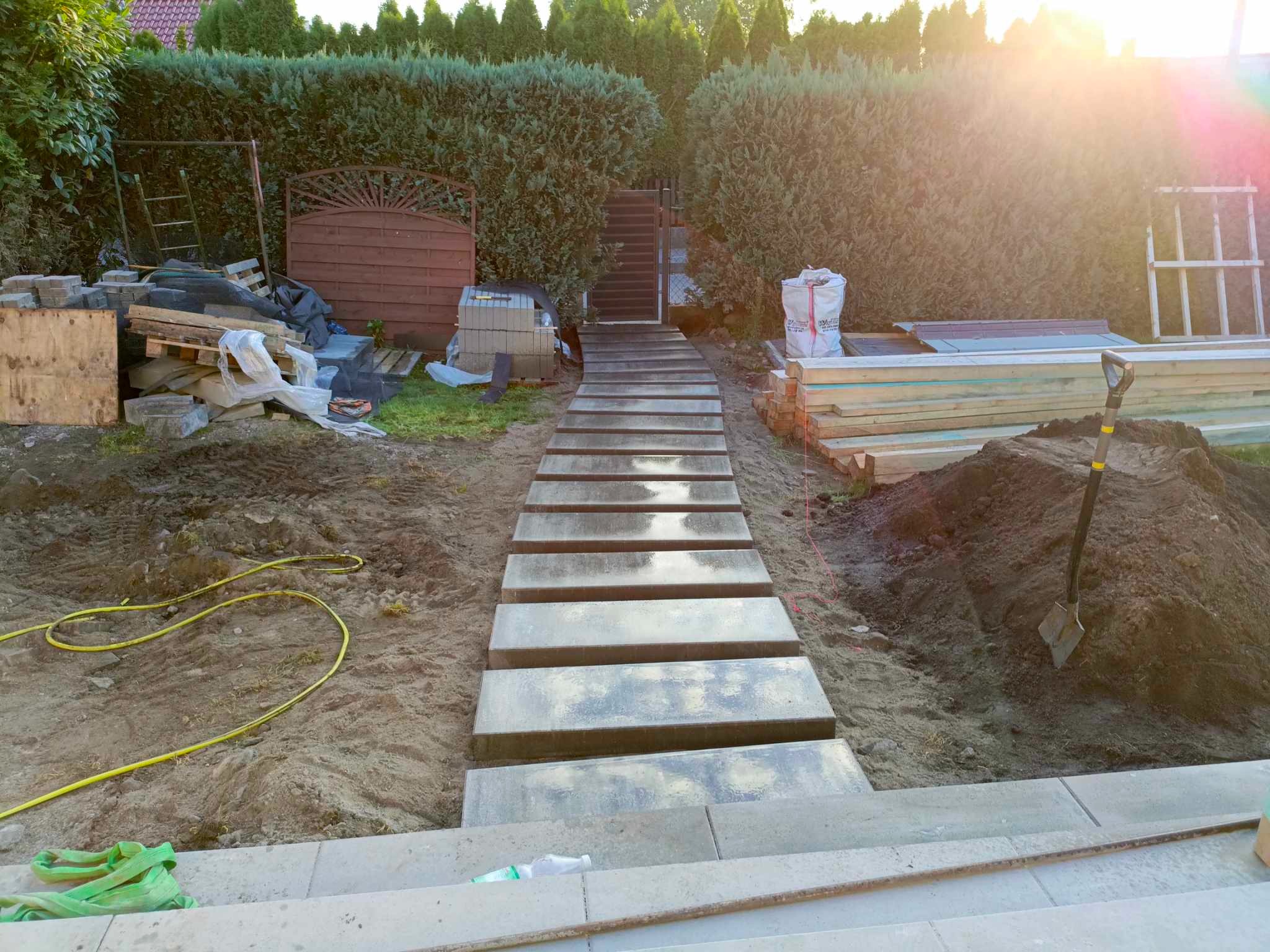 Ścieżka z betonowych płyt prowadząca do schodów w ogrodzie, otoczona ziemią i zielenią. Widoczne narzędzia i materiały budowlane. Słoneczne oświetlenie.