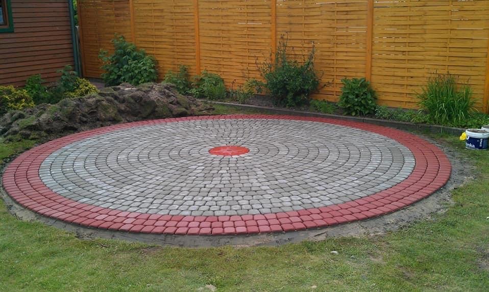 Okrągły plac z kostki brukowej w odcieniach szarości i czerwieni, z centralnym punktem w kolorze czerwonym. W tle drewniany płot i fragment budynku.