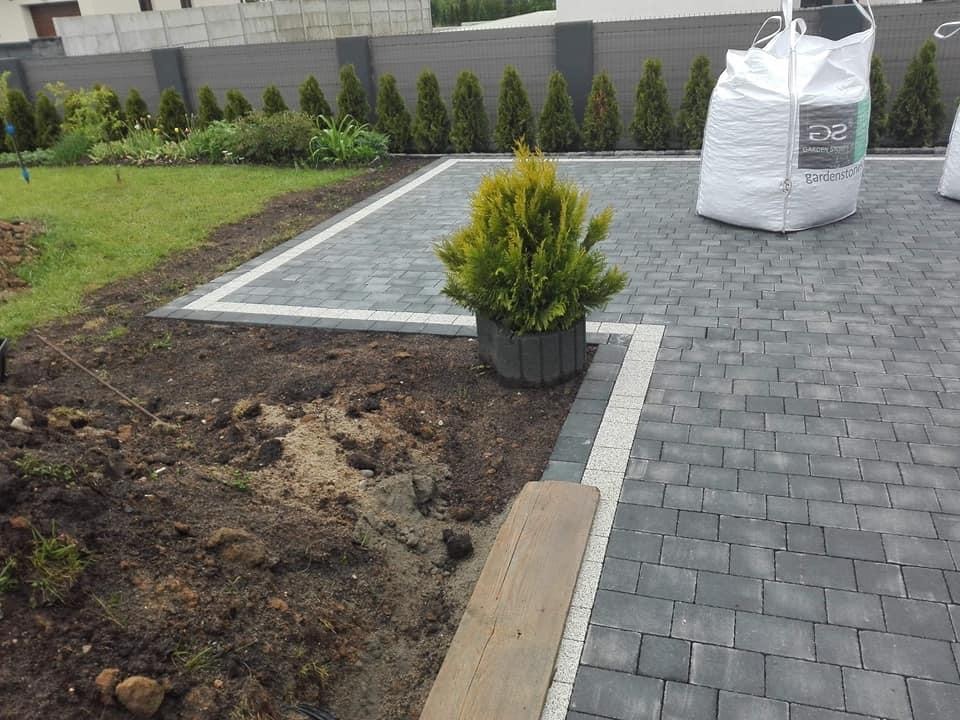 Ułożona szara kostka brukowa z białym obramowaniem, donica z iglakiem, worki z materiałem budowlanym w tle, trawnik i rząd tuj.