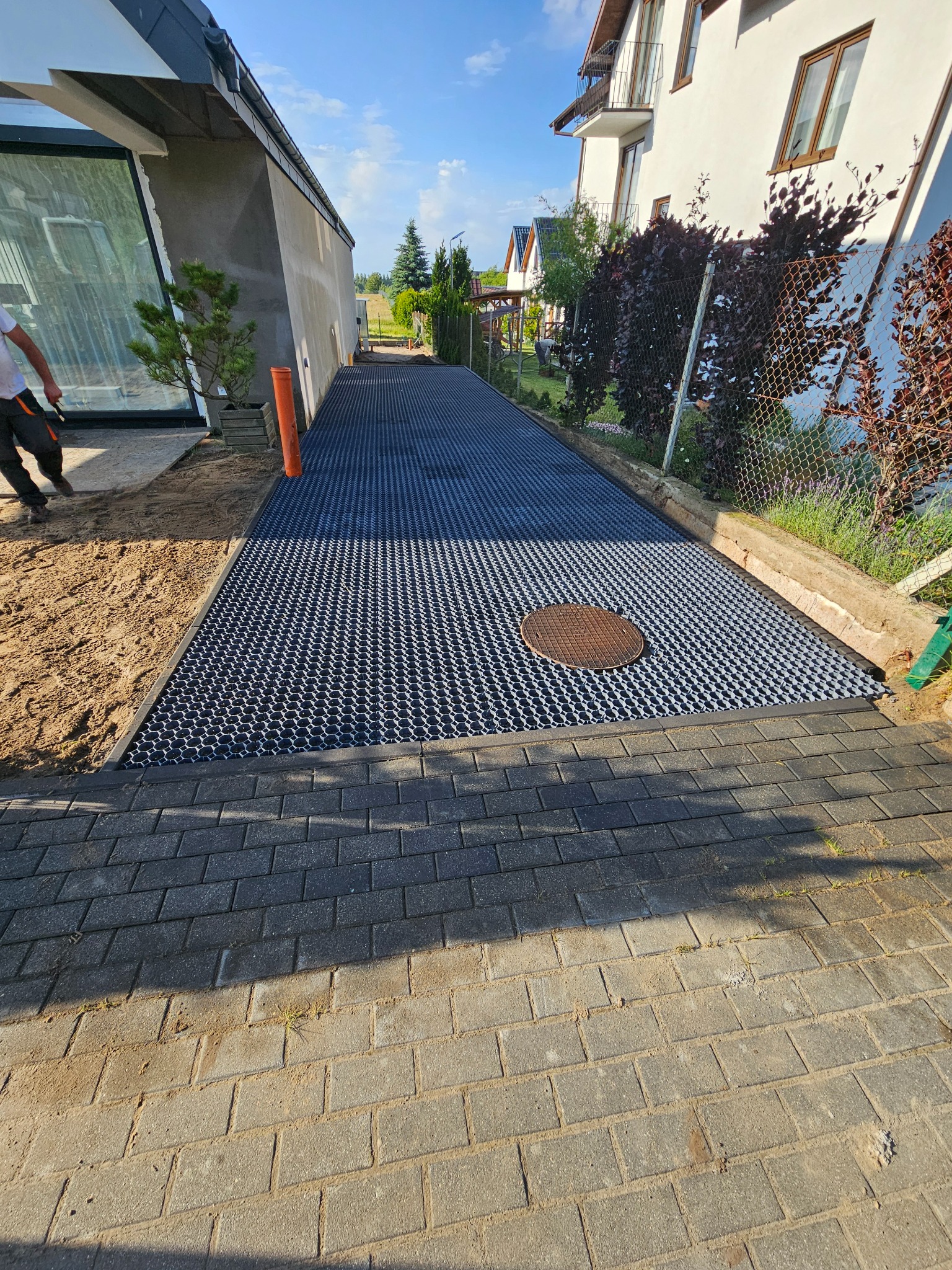 Ułożona geokrata na podbudowie z kostki brukowej, widoczny właz kanalizacyjny. W tle budynki mieszkalne i zieleń. Częściowo widoczna osoba w lewym górnym rogu.