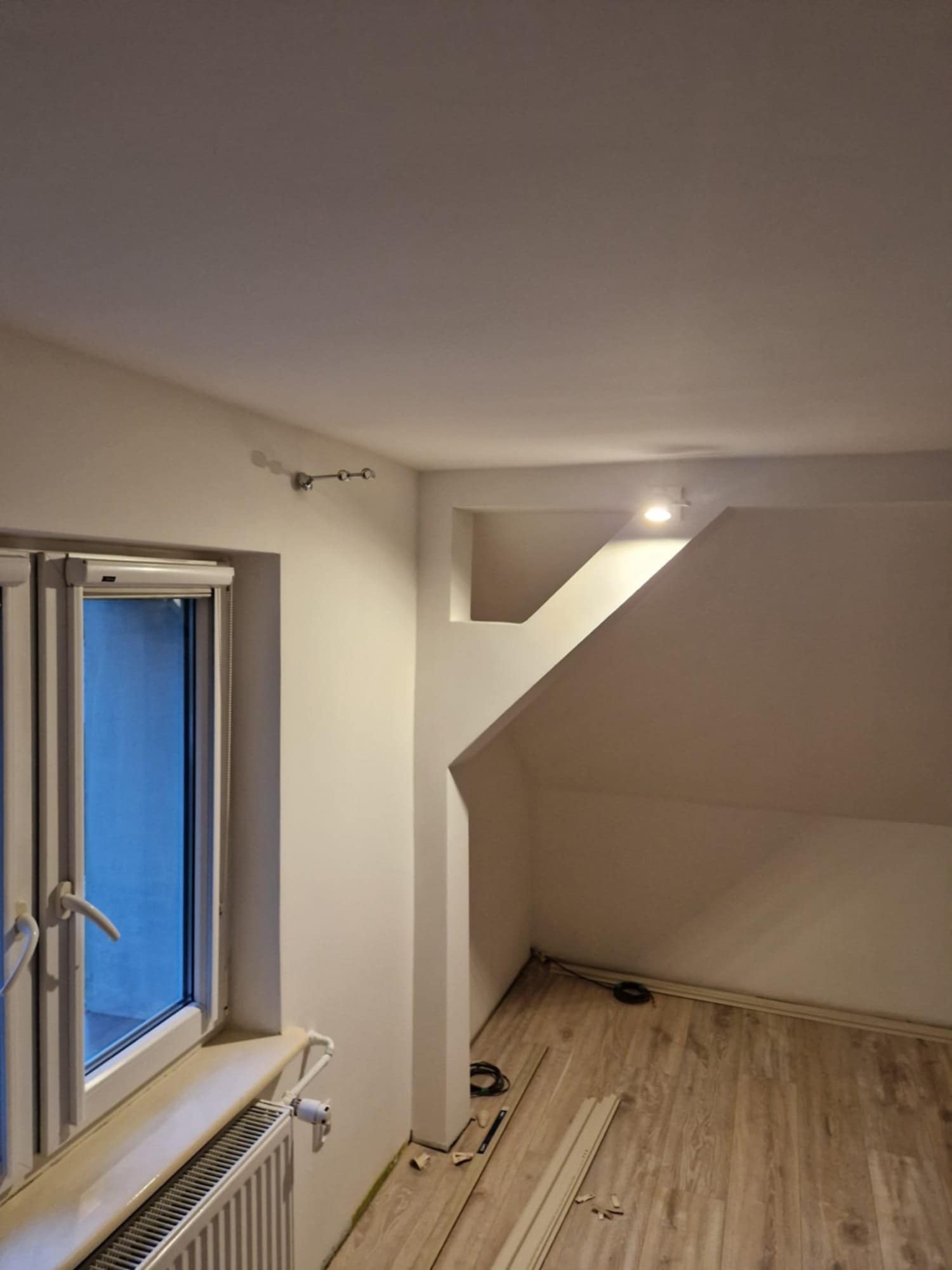 Jasne wnętrze poddasza z oknem, grzejnikiem i drewnopodobną podłogą. Widoczne elementy wykończeniowe i oświetlenie punktowe. Ściany pomalowane na biało.