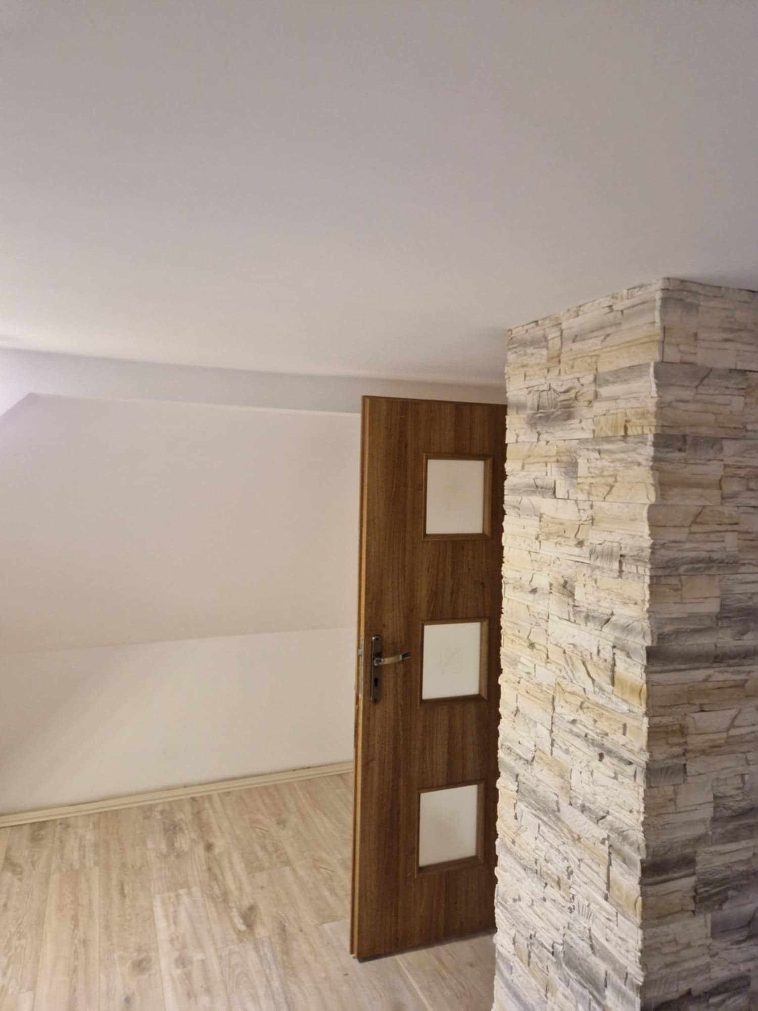 Wykończone wnętrze: drewniane drzwi z szybkami i dekoracyjny filar z kamienia w odcieniach szarości i beżu, na podłodze panele imitujące drewno, białe ściany i sufit.