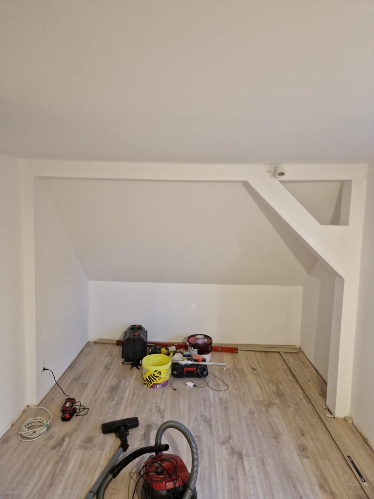 Pokój na poddaszu w trakcie prac wykończeniowych: panele podłogowe, białe ściany ze skosami, narzędzia i akcesoria budowlane. Widoczny odkurzacz.