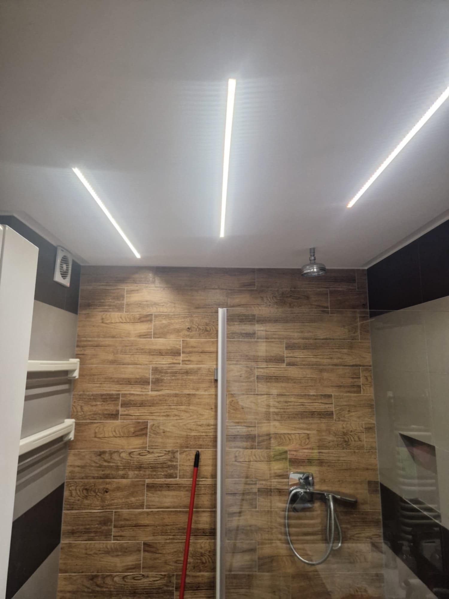 Nowoczesna łazienka z drewnopodobnymi płytkami i liniowym oświetleniem LED na suficie. Widoczna kabina prysznicowa i chromowana armatura.