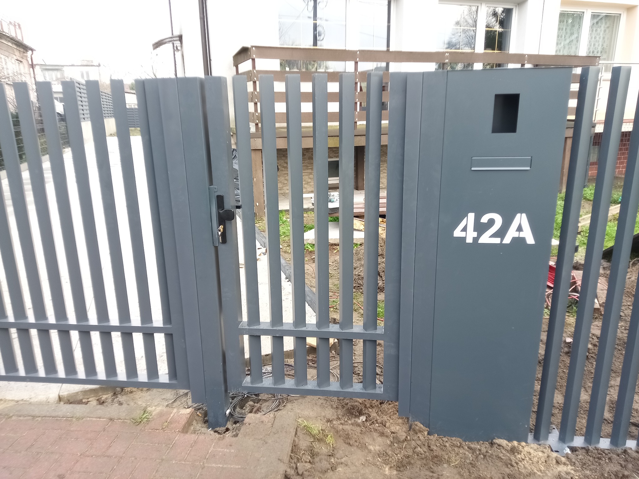 Nowoczesna, grafitowa brama wjazdowa z numerem domu 42A i skrzynką na listy. Pionowe, metalowe profile tworzą ażurową konstrukcję. Widoczne okablowanie przy podstawie.