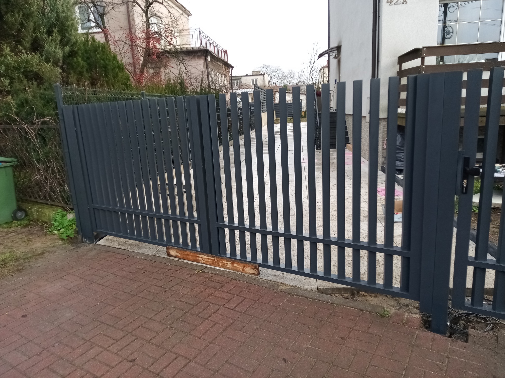 Nowoczesna, grafitowa brama wjazdowa z pionowych profili, widoczna z zewnątrz posesji na tle kostki brukowej i zabudowy miejskiej. Minimalistyczny design.