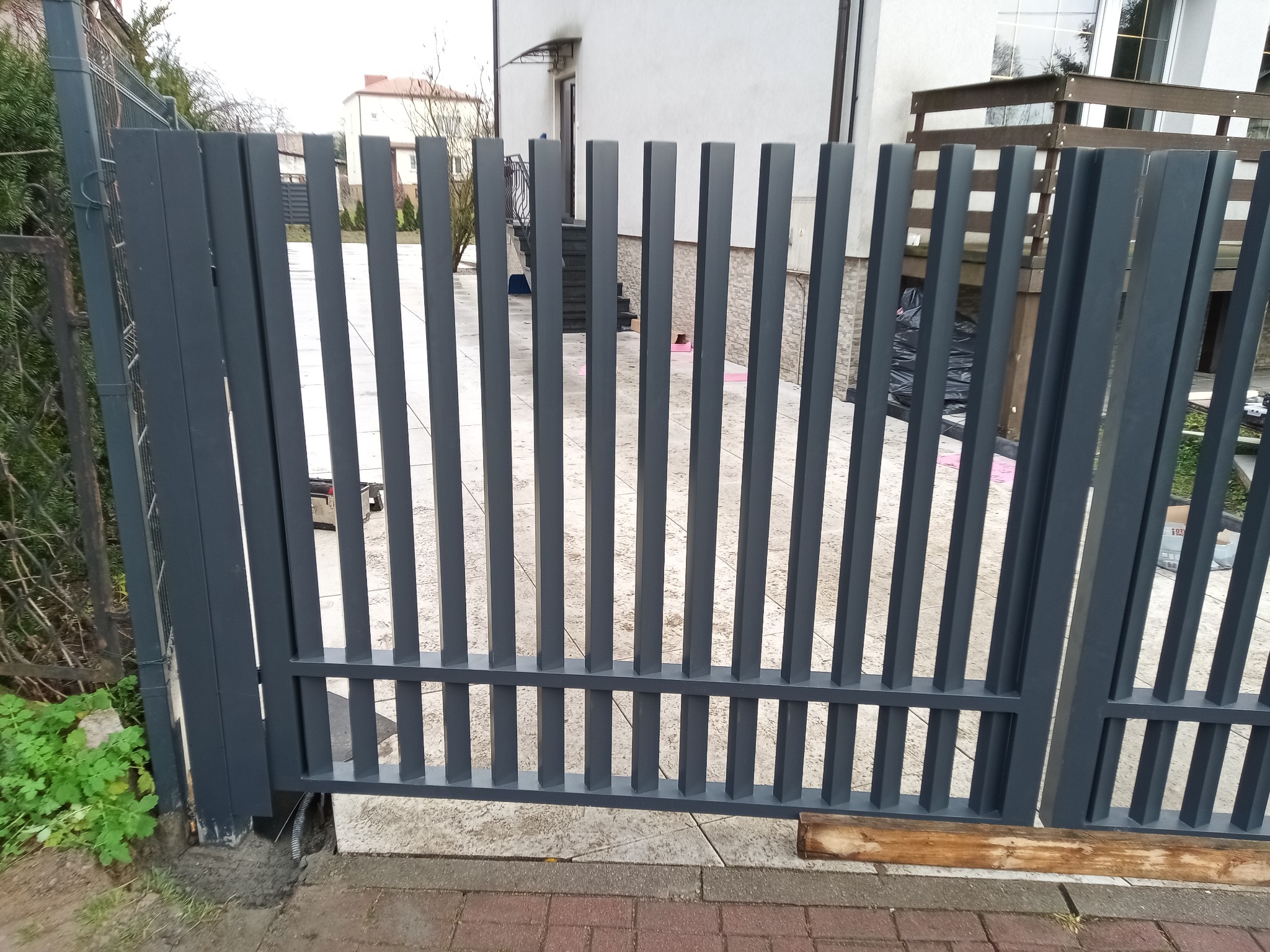 Nowoczesna, grafitowa brama wjazdowa z pionowych profili, widok z zewnątrz. Minimalistyczny design, jasne tło domu. Elementy montażowe widoczne u dołu.