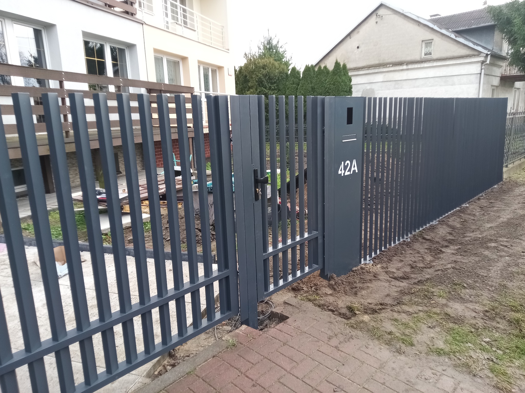 Nowoczesna, grafitowa brama z furtką i skrzynką na listy z numerem 42A. Pionowe, płaskie profile tworzą minimalistyczny design. Widok z zewnątrz posesji.