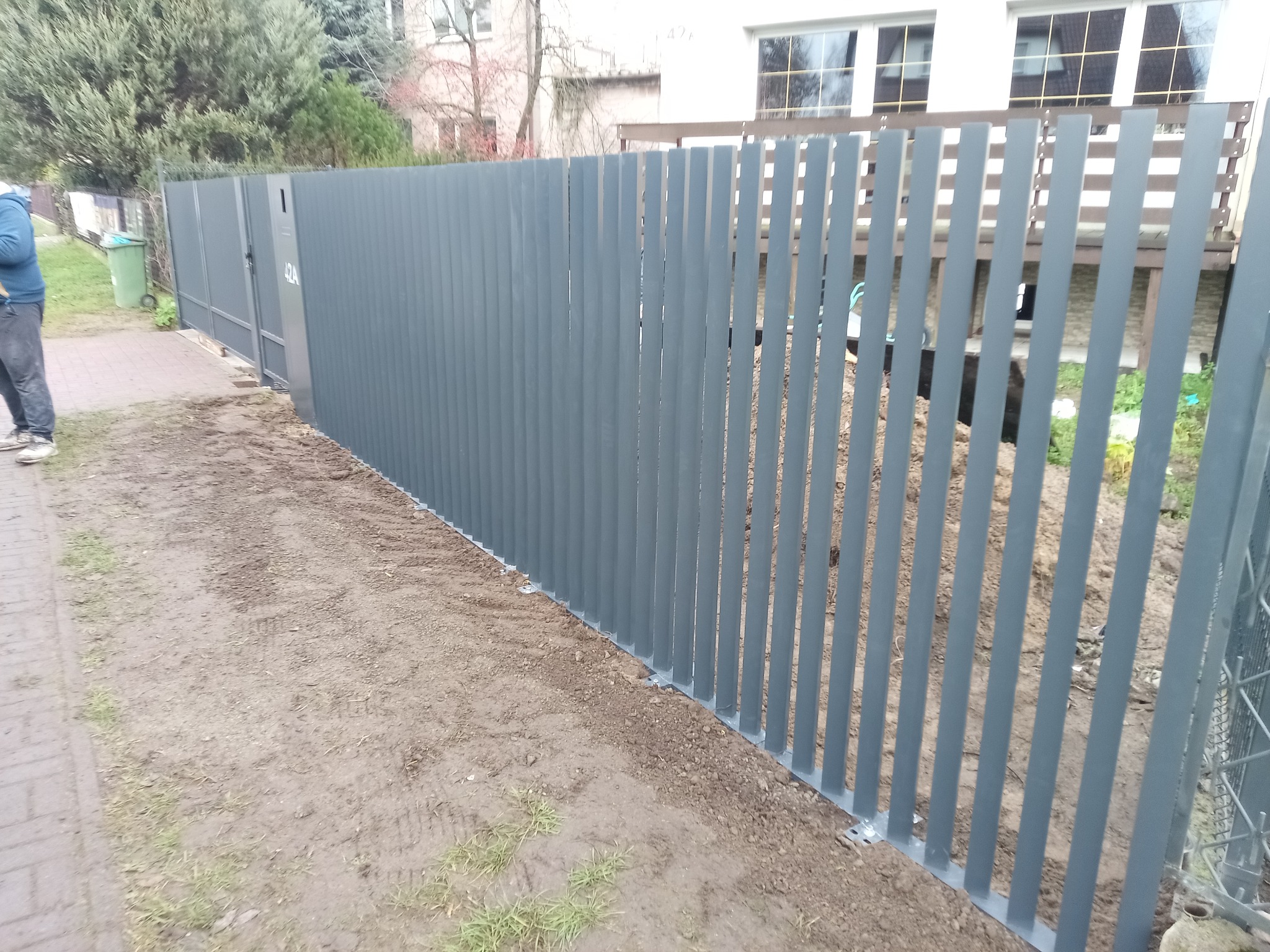 Nowoczesne, grafitowe ogrodzenie metalowe z pionowych profili, z bramą wjazdową z numerem 42A, na tle domu z białymi oknami. Widoczny fragment chodnika i trawnika.