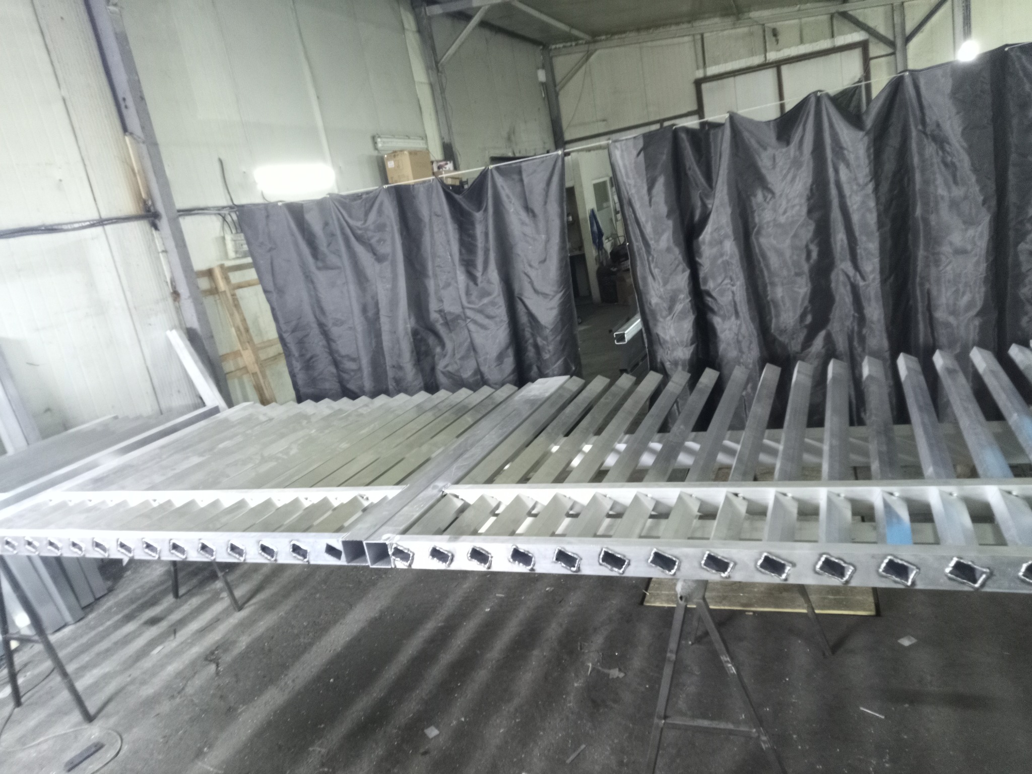 Metalowa konstrukcja z otworami, prawdopodobnie element ogrodzenia lub bramy, w warsztacie. Widoczne ślady spawania i szlifowania. W tle zasłona spawalnicza.