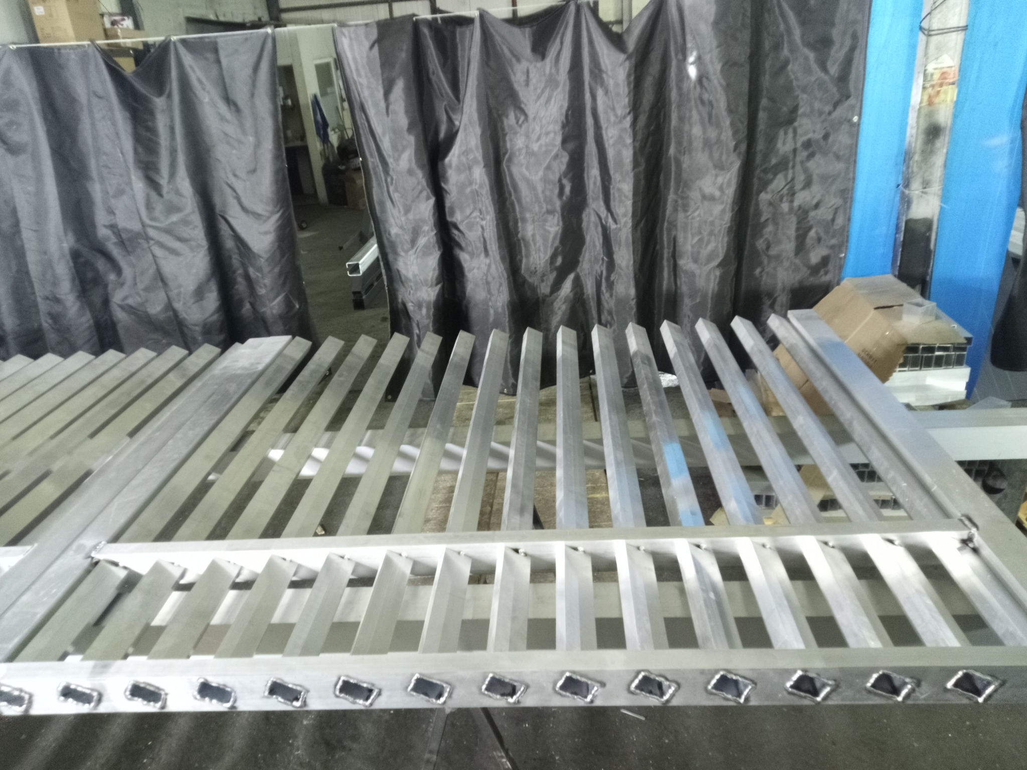 Surowa, aluminiowa konstrukcja bramy wjazdowej w warsztacie, widoczne spawy i elementy montażowe. Tło stanowią zasłony spawalnicze.