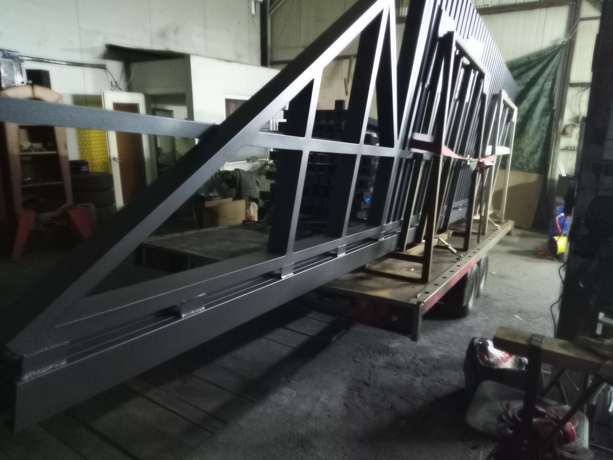 Szkielet metalowej bramy wjazdowej, pomalowany na szaro, umieszczony na przyczepie w warsztacie. Widoczne spawane elementy konstrukcyjne i fragmenty otoczenia warsztatowego.