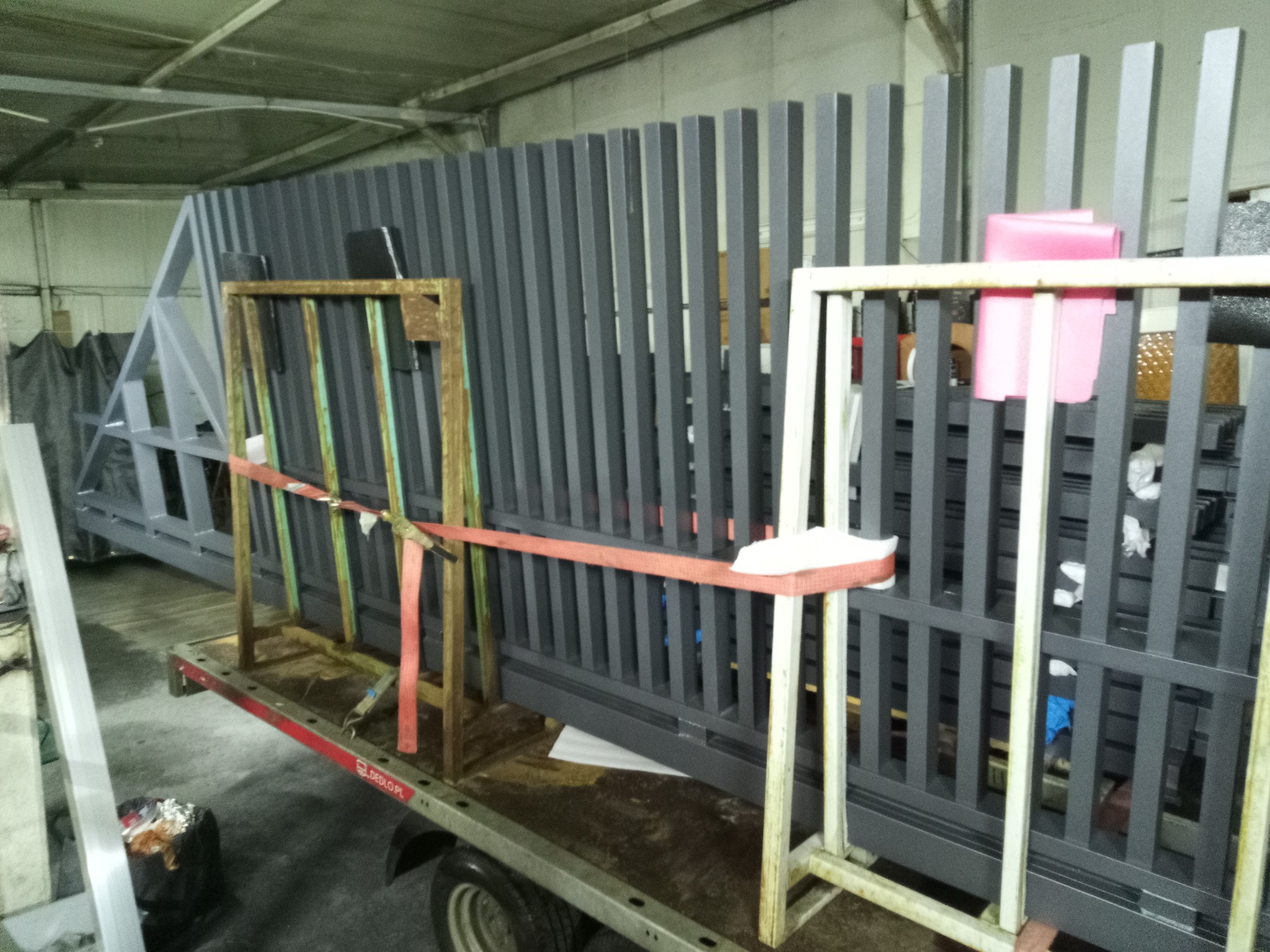 Szare, metalowe elementy konstrukcyjne, prawdopodobnie fragmenty bramy, zabezpieczone pasami na przyczepie w hali produkcyjnej. Widoczne detale spawów i łączeń.