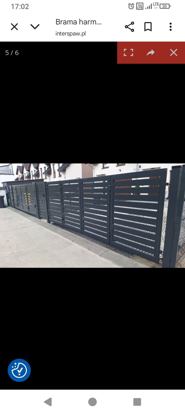 Nowoczesna, grafitowa brama przesuwna z poziomymi szczeblami, zamontowana przed domem z jasną elewacją. Widok z boku, w tle zieleń i fragment ogrodzenia.