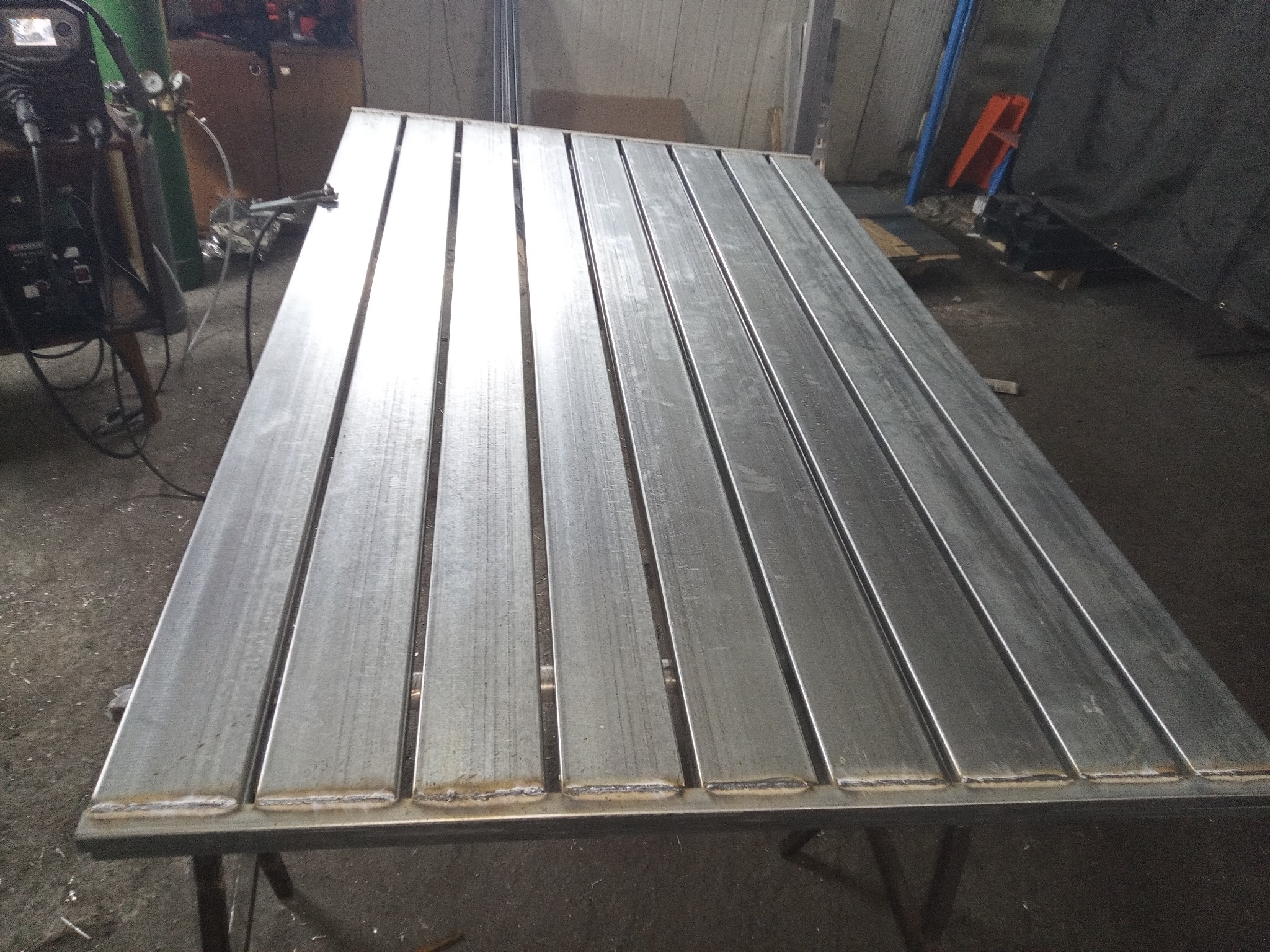 Stół spawalniczy z metalowych profili, widoczne spawy, w tle spawarka i butla z gazem. Konstrukcja warsztatowa z bliska, surowy metal.