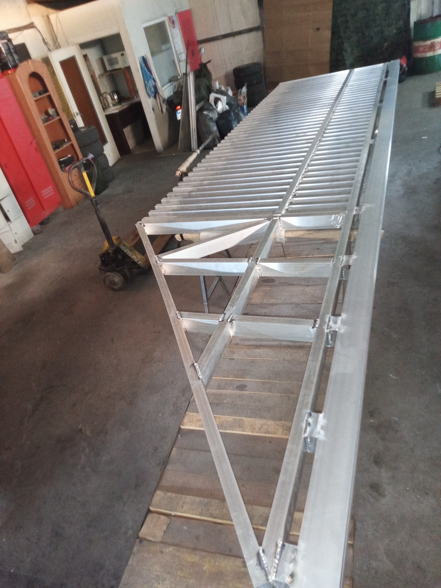 Konstrukcja metalowa z profili, prawdopodobnie rampa, w warsztacie. Widoczne spawy i elementy nośne. Ułożona na paletach, w tle wózek paletowy.