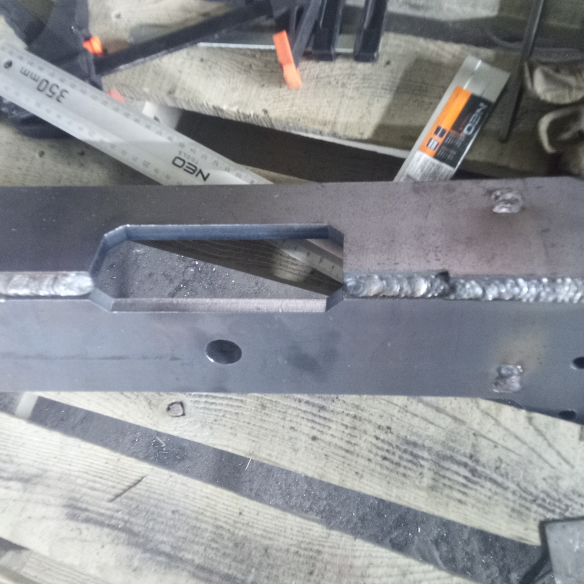 Metalowy element konstrukcyjny po spawaniu, widoczne spoiny, otwór i narzędzia pomiarowe na drewnianym podłożu. Detal obróbki metalu.