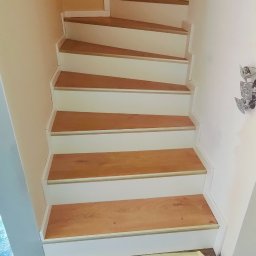 Bak-Pol Baka Krzysztof - Wykończone schody z drewnianymi stopniami i białymi podstopnicami. Widoczne detale wykończenia i zabezpieczona podłoga. Ujęcie z dołu schodów.