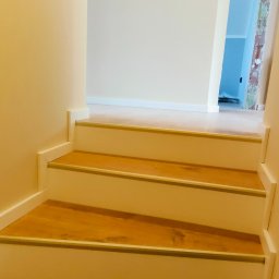 Bak-Pol Baka Krzysztof - Wykończone schody z drewnianymi stopniami i białymi podstopnicami, zabezpieczone metalowymi listwami. Widok z dołu, jasne ściany i fragment korytarza w tle.