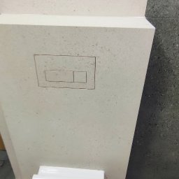 Bak-Pol Baka Krzysztof - Kompakt WC z zabudową z jasnego kamienia z przyciskiem spłukującym, na tle ściany z ciemnych płytek. Minimalistyczny design, nowoczesna łazienka.